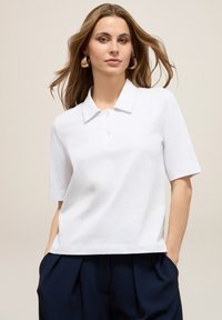 Chemise polo blanche avec un col classique, des manches courtes et une patte de boutonnage à trois boutons ; le tissu semble lisse et léger.