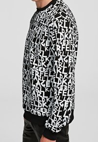 Svart sweatshirt med ett upprepande mönster av vitt logo. Rund hals, ribbade muddar och nederkant, texturerat tyg. Sidsyn som visar passformen.