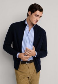 Marinefarbener Zip-Up Cardigan aus weichem Stoff, kombiniert mit einem hellblauen, gestreiften Hemd mit Knöpfen. Beigefarbene Cordhose und dunkler Gürtel im Detail.