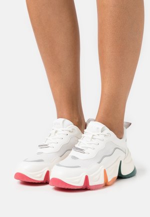 Baskets blanches épaisses avec semelles épaisses multicolores portées sur des jambes nues, avec des panneaux en mesh et cuir et des lacets noués.