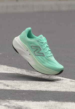 Chaussure de course New Balance vert clair en l’air au-dessus de l’asphalte avec des particules de saleté et des marquages routiers blancs.