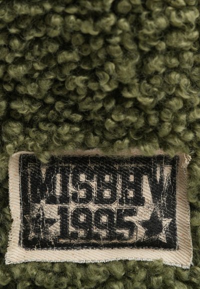 Olive green textured fabric with curly fibers, featuring a woven label in black and white that reads "MISBHV 1995."