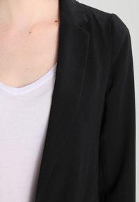 Černý blazer s módním střihem, lehkou tkaninou a klopami, vrstvený přes bílé tričko s výstřihem do V.