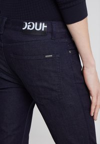 Mörkblå denimbyxor med en slät yta, försedda med en logotag, bakficka och subtila sömnadsdetaljer.