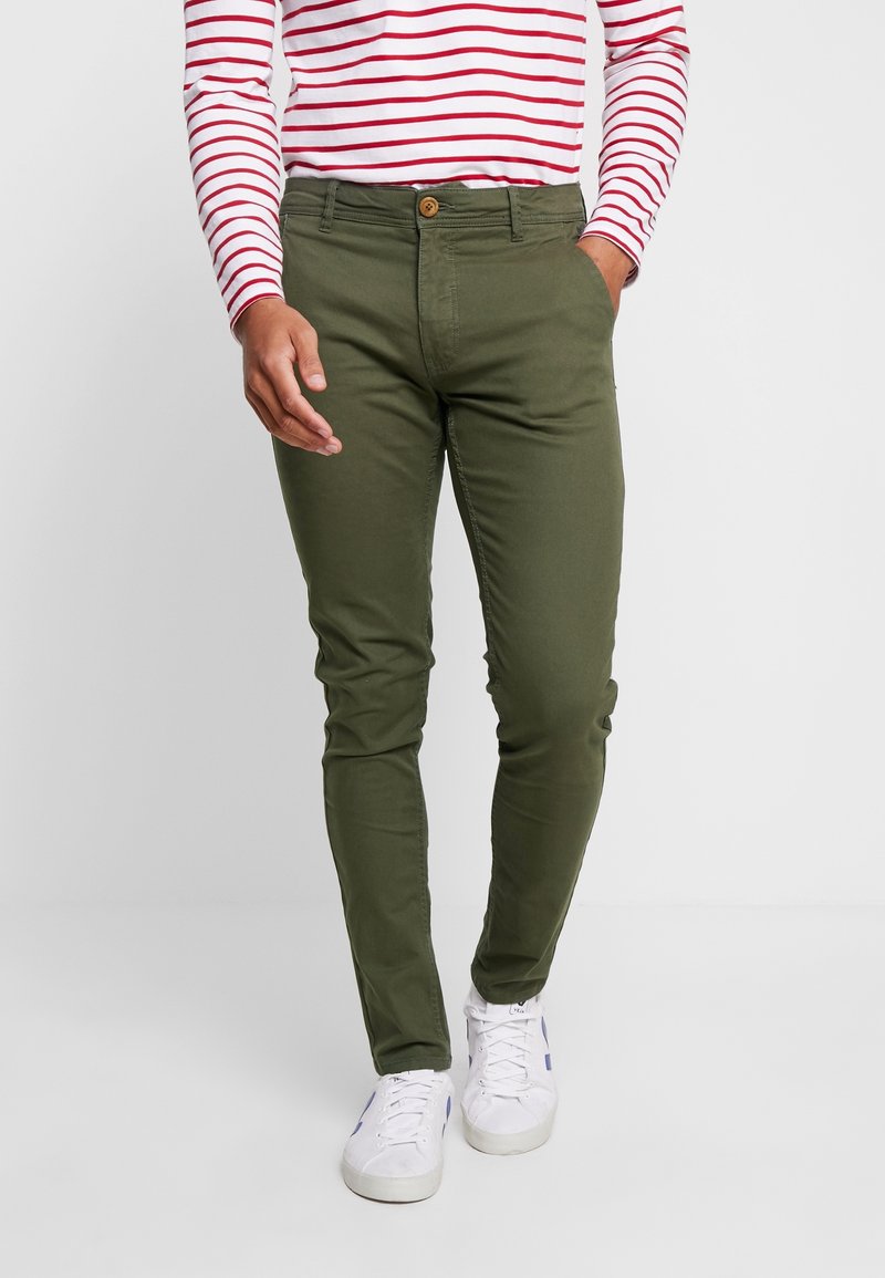 Homme portant un pantalon slim vert olive, des baskets blanches et une chemise à manches longues blanche avec des rayures horizontales rouges, debout avec une main dans la poche.