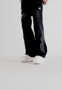 Pantalones de mezclilla negros con áreas desgastadas y bordado decorativo blanco a lo largo de los lados, combinados con zapatillas blancas gruesas.