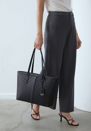 Personne portant un pantalon noir à jambes larges et un haut blanc, tenant un grand sac cabas noir à motifs et portant des sandales à talons noires avec lanières.