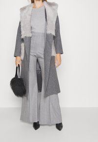 Woman wearing a long grey coat with light fur collar, grey pinstripe wide-leg pants, light grey top, black pointed boots, and holding a black furry handbag.