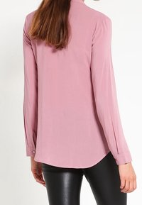 Blouse à manches longues, de couleur rose clair, avec une texture lisse, un ourlet incurvé et des poignets boutonnés, associée à un pantalon noir ajusté.