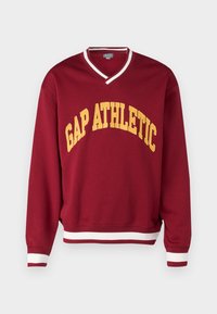 Sudadera burdeos con cuello en V, con un collar a rayas blancas y rojas, y "GAP ATHLETIC" en texto amarillo en arco. Material suave y puños acanalados.