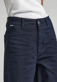 Tumši jūras zila džinsu bikses ar gludu tekstūru, piecu kabatu dizainu un metāla pogas aizdari. Uz jostas ir redzama nekonstatējama zīmola birka.