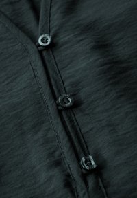 Tummanharmaa nappikauluspaita, jossa on teksturoitu kangas, neljä mustaa nappia ja siistit saumat läpässä ja kauluksessa.