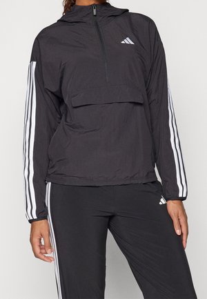 Henkilö pukeutuneena mustaan Adidas-verryttelypukuun, jossa valkoiset raidat hihansuissa ja lahkeissa, edessä vetoketju ja huppu, seisoo yksivärisen taustan edessä.