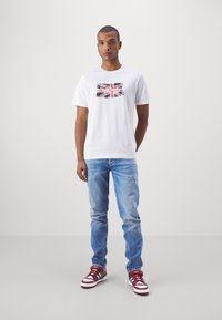 Witte katoenen t-shirt met een bedrukte Union Jack in rood, zwart en wit, gecombineerd met lichtblauwe jeans en rode sneakers.