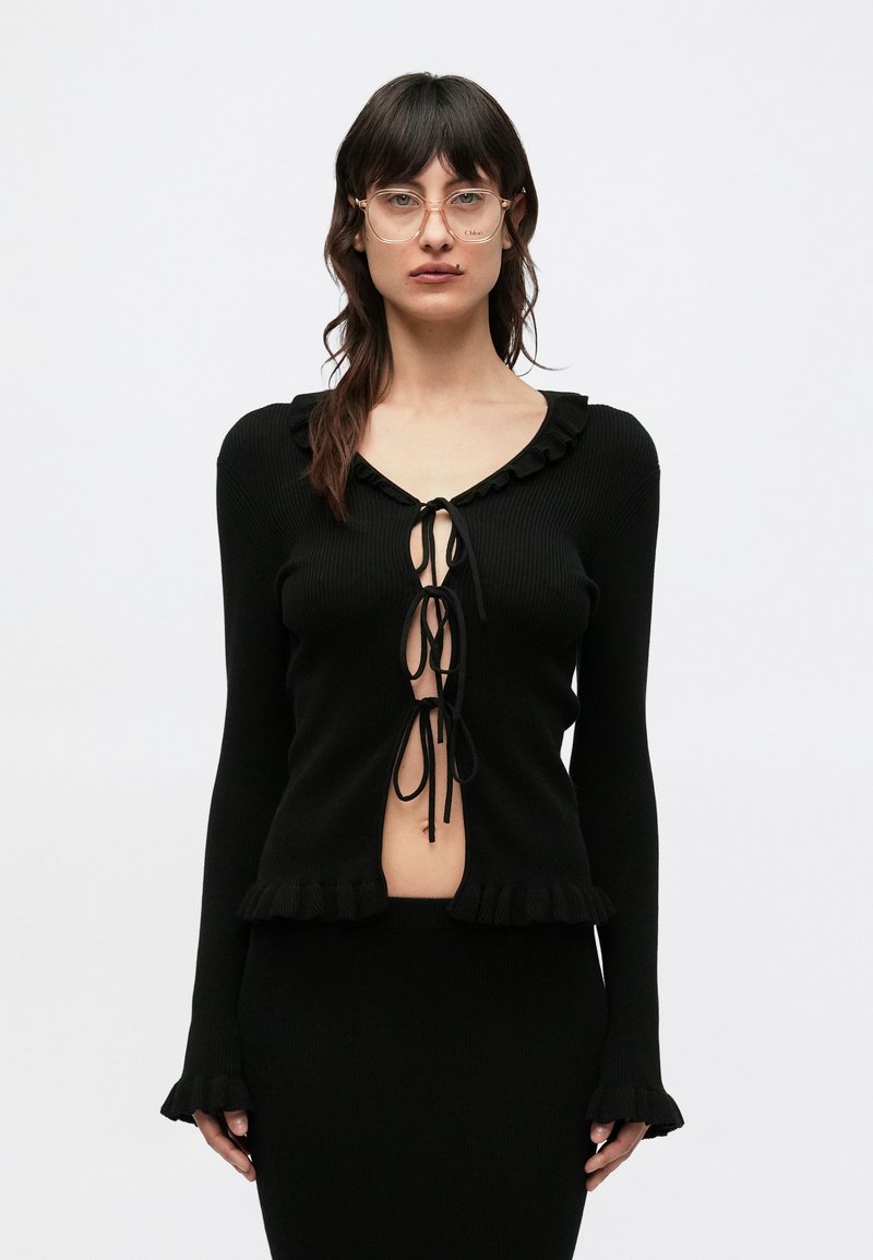 Mustast soonilisest kangast tumevoldiline pluus V-kaelusega, volangiliste servade ja keskel paeladega. Pikad varrukad lõdvendatud mansettidega, minimalistlik disain.