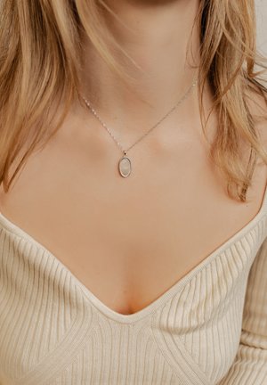 Silberne Halskette mit einem ovalen Anhänger, der eine strukturierte Oberfläche aufweist. Die Kette ist fein und zart und wird über einem gerippten, cremefarbenen Oberteil getragen.