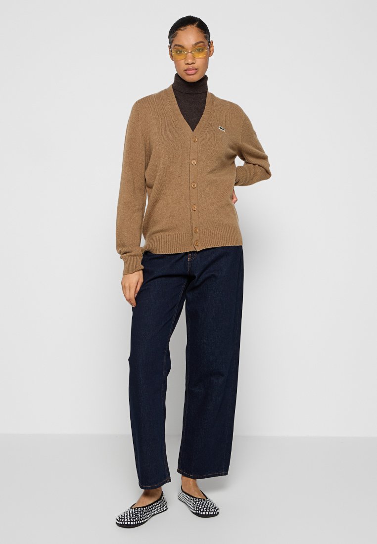 Cardigan marron avec un col en V et une fermeture boutonnée. Associe à un jean large bleu foncé et des chaussures slip-on à motifs. Tissu texturé.