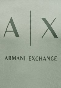 Λογότυπο A|X με κατακόρυφη γραμμή ανάμεσα στα γράμματα και το κείμενο «Armani Exchange» από κάτω, επάνω σε ανοιχτό πράσινο φόντο υφάσματος.