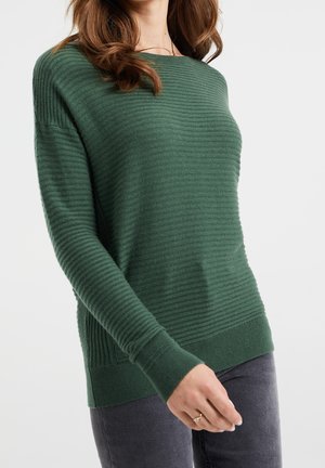 Vrouw met een geribbelde donkergroene trui met lange mouwen en donkergrijze broek, gekruld bruin haar en een ring aan haar linkerhand.