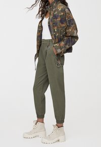 Veste camouflage avec motifs verts, orange et violets ; pantalon fuselé vert olive avec détail anneau ; bottes épaisses couleur crème.