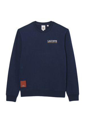 Sudadera azul marino de Lacoste con texto "Roland Garros Club" en el pecho y pequeño parche marrón con logo de cocodrilo cerca del dobladillo.