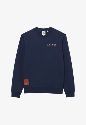 Sudadera azul marino de Lacoste con texto "Roland Garros Club" en el pecho y pequeño parche marrón con logo de cocodrilo cerca del dobladillo.