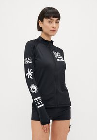 Črna dolgoročlenska rash guard s belimi grafičnimi odtisi, vključno z logotipom, palmami in valovi. S prednjim zadrgo in prilegnjenim dizajnom.