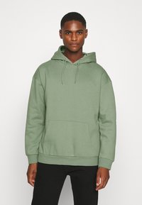 Light green hooded sweatshirt made of soft fabric, featuring a front pocket and ribbed cuffs, paired with black pants.