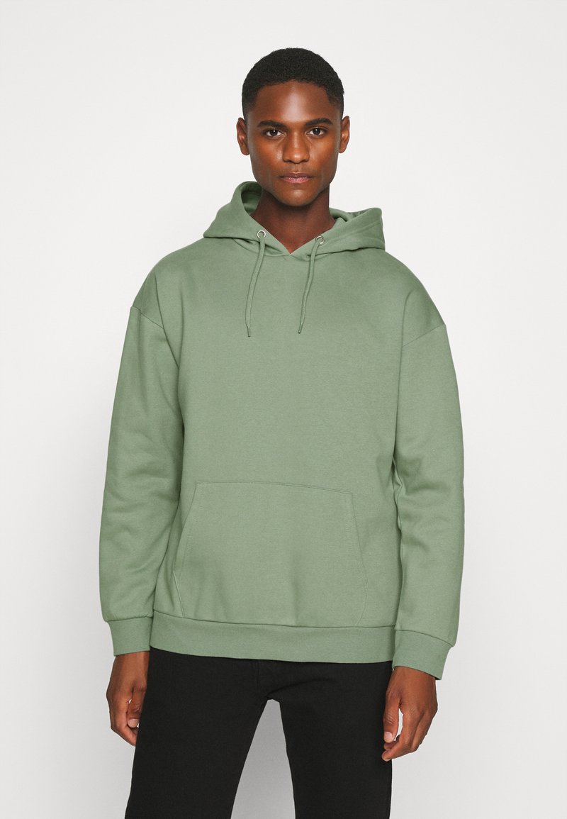 Light green hooded sweatshirt made of soft fabric, featuring a front pocket and ribbed cuffs, paired with black pants.