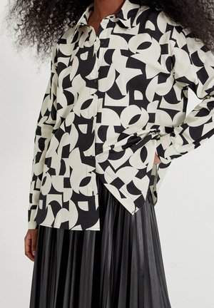 Black-and-white abstract patterned button-up shirt paired with a high-waisted black pleated skirt, model’s hand on hip.
