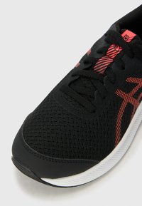 Baskets noires de sport avec une tige en mesh, des détails texturés, des accents rouges et une semelle blanche. Les lacets et le logo sont visibles sur le côté.