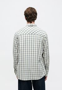 Mann iført en langermet hvit og blå rutete skjorte, vendt bort fra en enkel hvit bakgrunn.