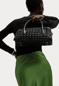 Schwarze handgefertigte Tasche mit einem Rautenmuster, glatten Akzenten, doppelten Griffen und Hardware-Details. Trägt man über der Schulter zu einem grünen Rock.
