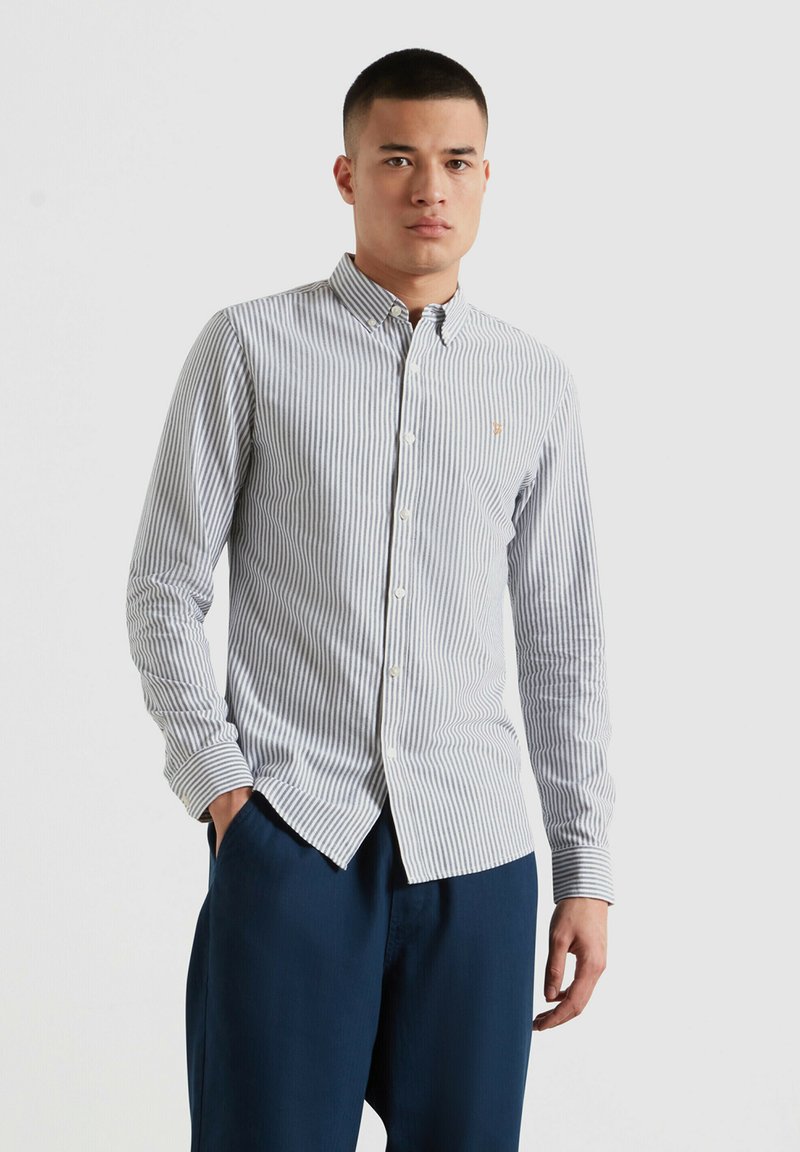 Jonge man die een overhemd met verticale strepen en donkerblauwe broek draagt, met één hand in de zak staand tegen een effen achtergrond.