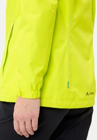 Veste imperméable jaune vif avec des poignets élastiques, des zips sur les côtés et un logo en bas à droite. Texture lisse, matériau léger.