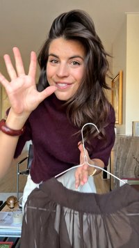 Dunkelbrauer Rock, der an einem weißen Kleiderbügel hängt, mit einem gerafften Bund und transparenter Stoffqualität. Eine Person hält den Bügel und lächelt.