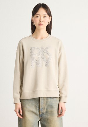 Beige genser med "DKNY"-logo laget av rhinestones, rund halslinning og ribbestrikkede ermer. Brukt over lyseblå slitte jeans.