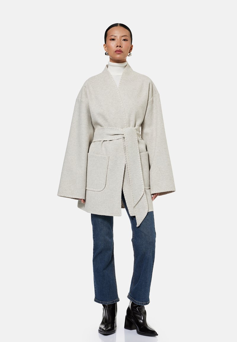Femme portant un manteau style kimono beige clair ceinturé, un pull à col roulé blanc, un jean bleu droit et des bottines noires à talons, debout immobile.