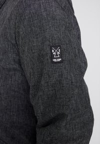 Chaqueta negra texturizada con un diseño ajustado y un parche de logo rectangular en la manga izquierda con gráficos de cruces y texto.