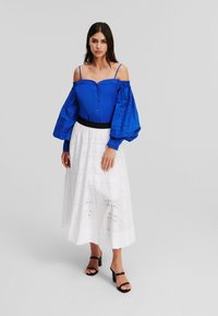 Blusa blu senza spalle con dettagli a bottoni e maniche voluminose, abbinata a una gonna bianca a livelli con ricami e sandali neri con tacco a blocco.