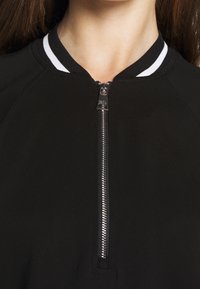 Close-up of a person wearing a black zip-up top with a silver zipper and a black collar featuring white stripes.