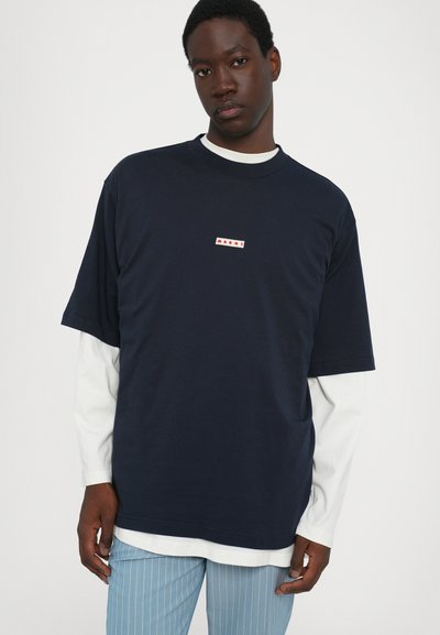 Mörkblå bomulls t-shirt med en liten vit/röd logo. Bärs över en långärmad vit skjorta, i kombination med ljusblå randiga byxor.