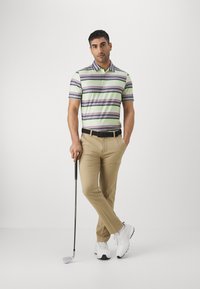 Camisa polo de hombre de manga corta con rayas verdes y moradas, combinada con pantalones beige y zapatos de golf blancos, sosteniendo un palo de golf.
