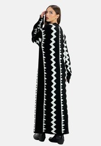 Robe longue noire avec motif zigzag blanc, texture tricotée, manches larges et coupe droite, portée avec des bottines jusqu'à la cheville.