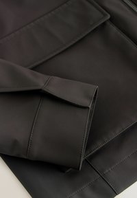Manche de veste noire avec poignet et couture détaillée sur tissu lisse, repliée sur le corps de la veste assortie avec fermeture éclair visible.