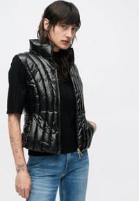 Čierna prešívaná vesta s vysokým golierom, lesklý materiál, zapínanie na zips a elastický lem. Nosená cez čierny sveter a kombinovaná s modrými džínsy.