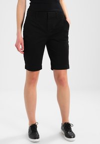Shorts en coton noirs à coupe droite, avec poches latérales et taille plate. Portés avec des baskets noires et un haut noir.