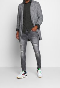 Grijze lange jas over een donker overhemd met knopen, gecombineerd met gescheurde grijze spijkerbroek en witte sneakers met meerkleurige accenten.