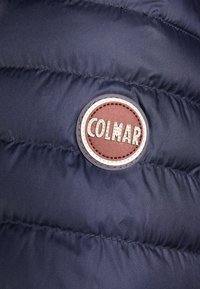 Marinblå pufferjacka med quiltad textur. Utrustad med en cirkulär brun Colmar-logga med vit sömnad och en yttre vit kant.