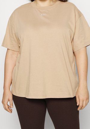 Osoba ubrana w luźny beżowy T-shirt z krótkim rękawem z małym białym logo przy dekolcie oraz ciemne, prążkowane legginsy, stojąca z rozluźnionymi ramionami.